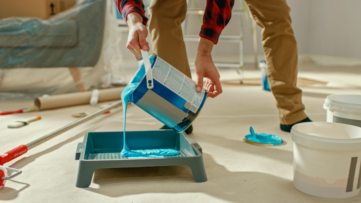 A man in khakis and a black and red plaid shirt pouring blue paint into a tray