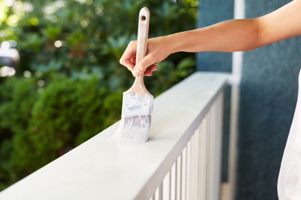 woman's hand holding a paintbrush as she brushes it across a deck railing, painting it white